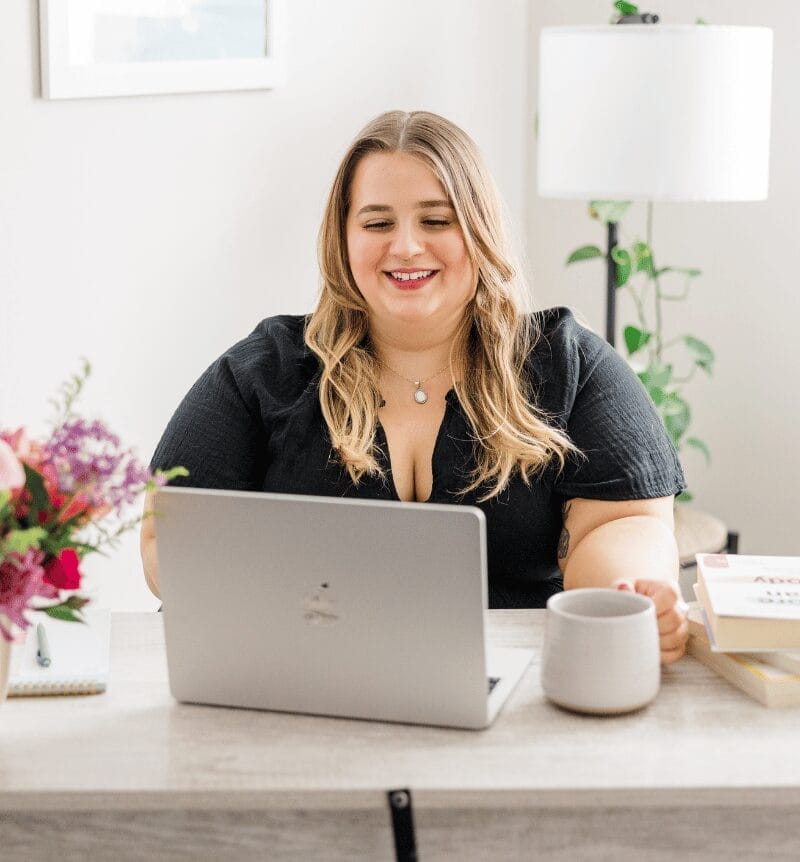 Woman in a black dress smiling at a laptop on a desk, holding a mug. Surroundings include books, a bouquet of flowers, and a cozy room ambiance.